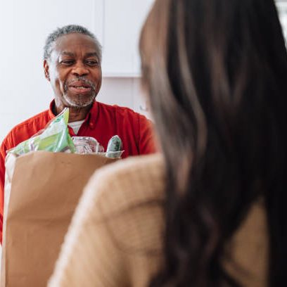 A young female Caucasian volunteer brings bag with food to an older man's front door